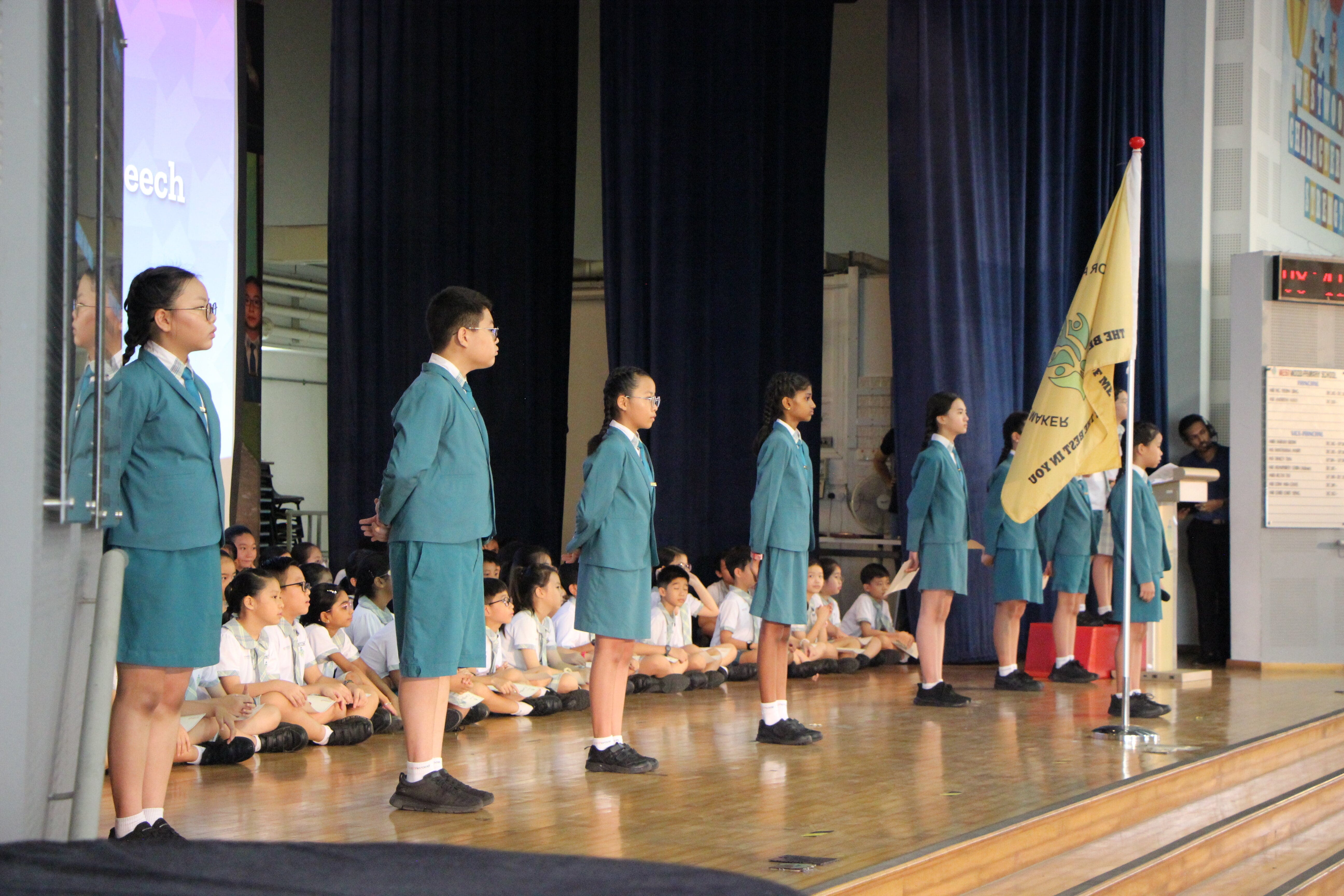 Students in teal uniforms stand in a line on a school hall stage beside a yellow flag, with seated pupils watching.
